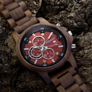 May include: A wooden wristwatch with a dark brown finish and a red watch face. The watch features three sub-dials and a matching wooden band. The watch is displayed on a textured, dark brown surface.