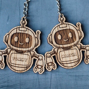 Può includere: Due portachiavi a forma di robot in legno con dettagli incisi. Ogni robot ha una faccia sorridente, un'antenna e i nomi "Tommy" e "Martha" sul petto. Sono attaccati a catene d'argento.