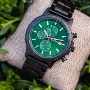 Op de afbeelding: Een donkerhouten polshorloge met een groene wijzerplaat en zwarte accenten. Het horloge heeft drie sub-wijzerplaten en een datumweergave. Het horloge ligt op een beige ondergrond, omringd door groen gebladerte. De horlogeband is van donker hout.