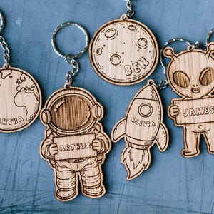 Könnte beinhalten: Holz-Schlüsselanhänger mit Weltraum-Motiven. Enthält einen Planeten Erde, einen Astronauten, einen Mond, eine Rakete und einen Außerirdischen. Jeder Schlüsselanhänger hat einen eingravierten Namen: Samantha, Arthur, Ben, Olivia und James. Die Schlüsselanhänger sind an silbernen Metall-Schlüsselringen befestigt.