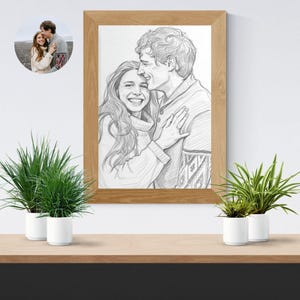May include: A framed pencil sketch of a couple embracing, with a smaller circular photo in the upper left corner. The sketch is in a wooden frame and features a man and woman in sweaters. Two potted plants sit on a wooden shelf below.
