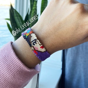 Puede incluir: Un brazalete de cuentas moradas y blancas con una imagen colorida de cuentas de un rostro de mujer. El brazalete está atado con un cordón negro. El texto "@ulv.creaciones" es visible en la muñeca.