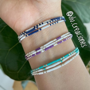 May include: Three layered bracelets with white, blue, purple, and turquoise beads. The bracelets are stacked on a person's wrist.