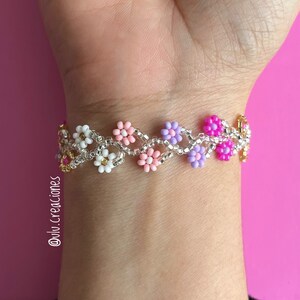 Puede incluir: Un brazalete de cuentas con una cadena plateada y cuentas en forma de flor de color rosa, blanco y morado. El brazalete está en la muñeca de una persona.