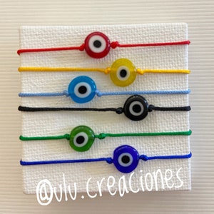 May include: Six colourful string bracelets with evil eye charms. The charms are red, yellow, blue, black, green, and blue. The bracelets are on a white background. The text '@viv.creaciones' is at the bottom of the image.