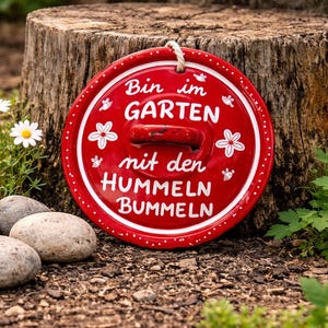 Könnte beinhalten: Rundes, rotes Gartenschild mit weißem Text und floralen Akzenten. Auf dem Schild steht "Bin im GARTEN mit den HUMMELN BUMMELN". Es hat einen Griff und hängt an einem weißen Seil. Das Schild steht vor einem Baumstumpf.
