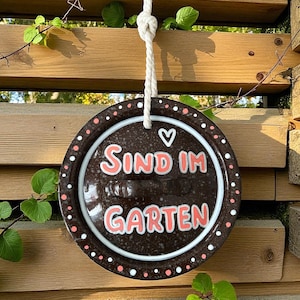 Könnte beinhalten: Rundes, braunes Keramik-Gartenschild mit den Worten "SIND IM GARTEN" in rosa Schrift. Das Schild ist mit weißen und rosa Punkten und einem weißen Herz verziert. Es hängt an einem weißen Seil vor einem Holzzaun.