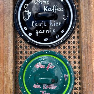 Könnte beinhalten: Zwei runde Keramikdeckel mit Griffen. Der obere Deckel ist schwarz mit weißen Punkten, Kaffeebohnen-Illustrationen und dem Text "Ohne Kaffee läuft hier garnix". Der untere Deckel ist grün mit weißen und rosa Punkten, einem Herz und dem Text "Was für ein toller Tag".