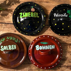Könnte beinhalten: Vier runde Topfdeckel mit Griffen. Zwei schwarze Deckel mit gelben Punkten und den Wörtern "Zwiebel" und "Petersilie". Die anderen beiden Deckel sind braun und rot, mit den Wörtern "Salbei" und "Bohnen".
