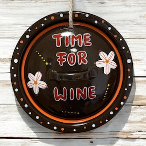 Könnte beinhalten: Runder, brauner Metallschild mit den Worten "TIME FOR WINE" in roter und weißer Schrift. Das Schild hat einen orangefarbenen Rand und ist mit weißen Punkten und Blumendesigns verziert. Ein Juteseil ist zum Aufhängen befestigt.