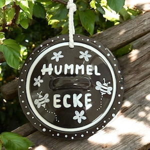 Könnte beinhalten: Ein braunes Keramik Schild mit weißen Punkten und einem weißen Rand. Das Schild trägt die Aufschrift "Hummel Ecke" in weiß mit einer stilisierten Bienenillustration auf jeder Seite des Textes. Es gibt auch stilisierte weiße Blumenillustrationen um den Text herum.