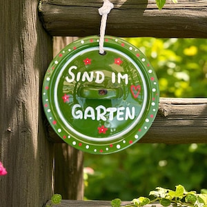 Könnte beinhalten: Runder, grüner Gartenschild mit dem Schriftzug "Sind im Garten" in Weiß. Verziert mit roten Blumen und weißen Punkten, hängt es an einem Holzzaun.