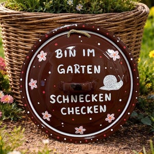 Könnte beinhalten: Ein runder, brauner Garten-Teller mit dem Text "BIN IM GARTEN SCHNECKEN CHECKEN" und einer Schnecken-Illustration. Der Teller hat einen weißen Rand mit roten Punkten und Blumenmotiven. Im Hintergrund ein geflochtener Blumenkorb.