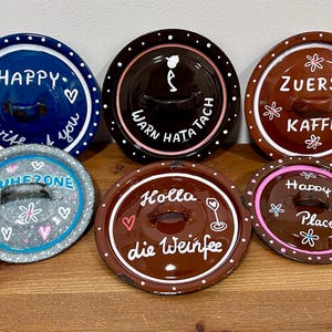Könnte beinhalten: Sechs Keramik-Topfdeckel mit handgemalten Designs und Text. Die Deckel sind in verschiedenen Farben gehalten, darunter Blau, Braun und Grau, mit weißen Punkten und dekorativen Akzenten. Der Text enthält "HAPPY", "ZUERST KAFFEE" und "Happy Place".