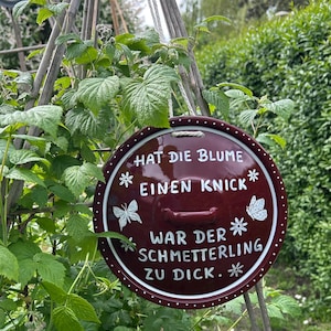 Könnte beinhalten: Ein runder, weinroter Gartenanhänger mit weißem Text und Illustrationen von Schmetterlingen und Blumen. Der Anhänger trägt die Aufschrift "HAT DIE BLUME EINEN KNICK WAR DER SCHMETTERLING ZU DICK." Der Anhänger ist von grünem Laub umgeben.