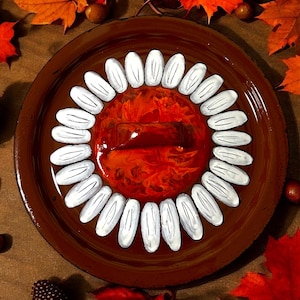 Könnte beinhalten: Ein brauner Keramikdeckel mit einem weißen Gänseblümchen-Design. Die Mitte des Gänseblümchens ist ein Wirbel aus orangefarbener und roter Farbe. Der Deckel hat einen braunen Griff.