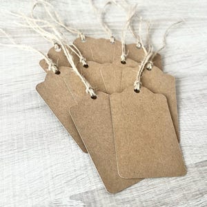 May include: A collection of blank, brown paper tags with scalloped edges and twine attached. The tags are rectangular with a rounded top and a hole for the twine. They are arranged on a light grey wooden surface.