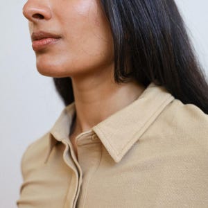 Pode incluir: Um close-up de uma mulher vestindo uma camisa bege de colarinho. A camisa tem um fecho de bot&otilde;es na frente e mangas compridas.