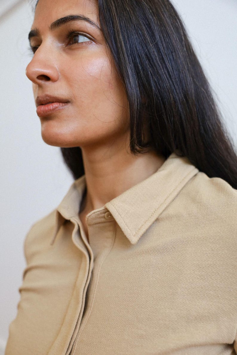 Pode incluir: Um close-up de uma mulher vestindo uma camisa bege de colarinho. A camisa tem um fecho de bot&otilde;es na frente e mangas compridas.