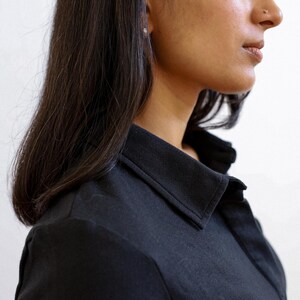 Pode incluir: Uma mulher com cabelo comprido e preto vestindo uma camisa preta com colarinho. A camisa tem um design simples e um caimento cl&aacute;ssico.