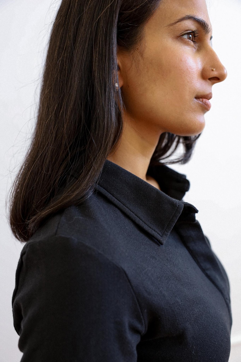 Pode incluir: Uma mulher com cabelo comprido e preto vestindo uma camisa preta com colarinho. A camisa tem um design simples e um caimento cl&aacute;ssico.