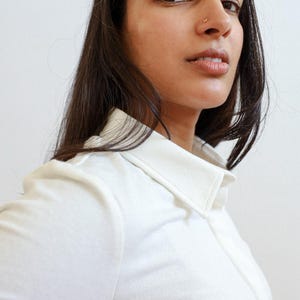Pode incluir: Uma mulher com cabelo comprido e escuro est&aacute; vestindo uma camisa branca de colarinho. Ela est&aacute; olhando diretamente para a c&acirc;mera.