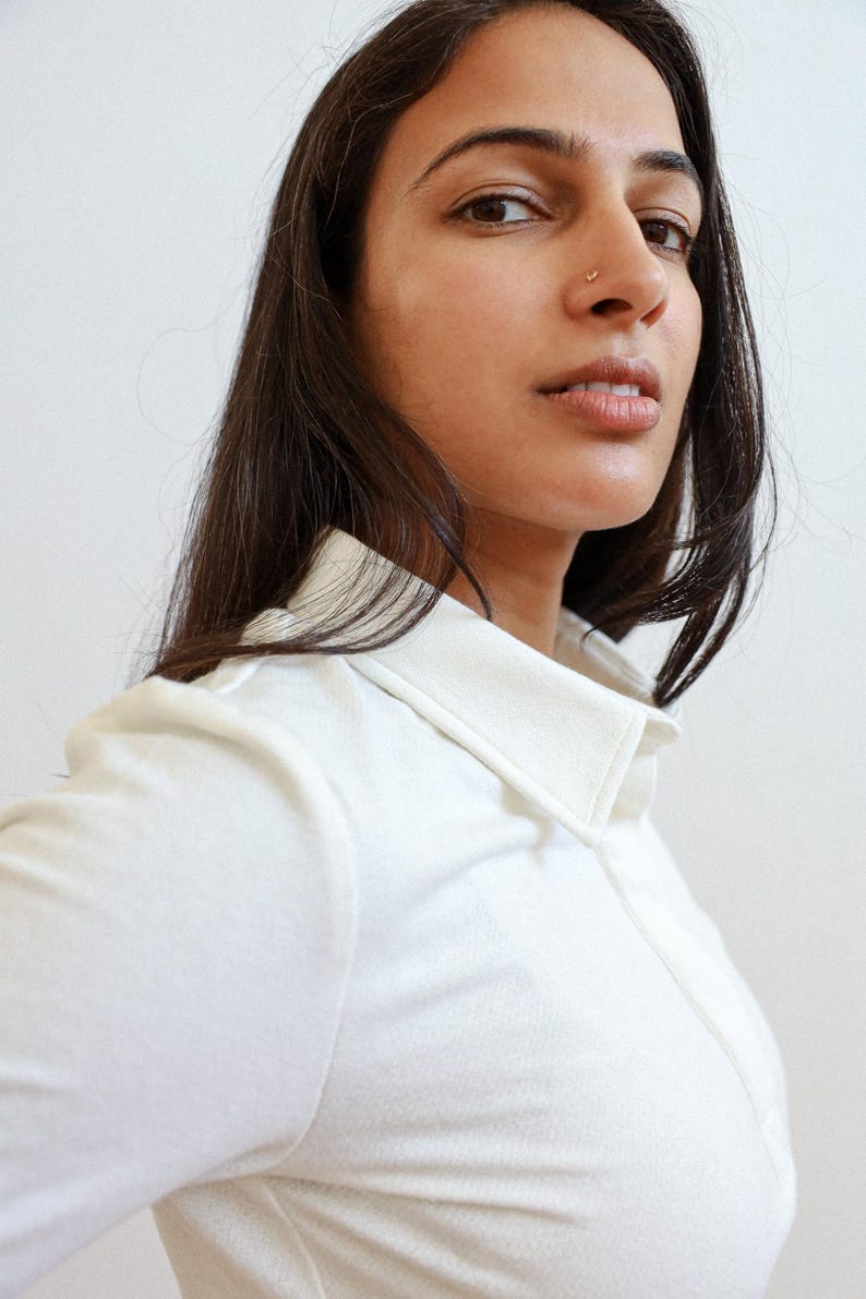 Pode incluir: Uma mulher com cabelo comprido e escuro est&aacute; vestindo uma camisa branca de colarinho. Ela est&aacute; olhando diretamente para a c&acirc;mera.