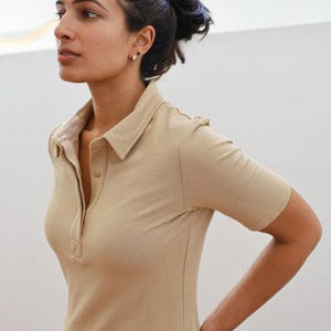 Pode incluir: Uma mulher vestindo uma camisa de manga curta com colarinho de cor marrom claro. A camisa tem um fecho de bot&otilde;es na frente e um caimento relaxado.