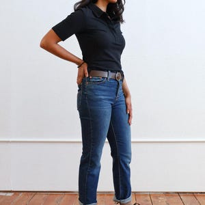 Pode incluir: Uma pessoa vestindo uma camisa preta de colarinho, jeans azul e botas de tornozelo marrons. A pessoa est&aacute; em p&eacute; em um piso de madeira.
