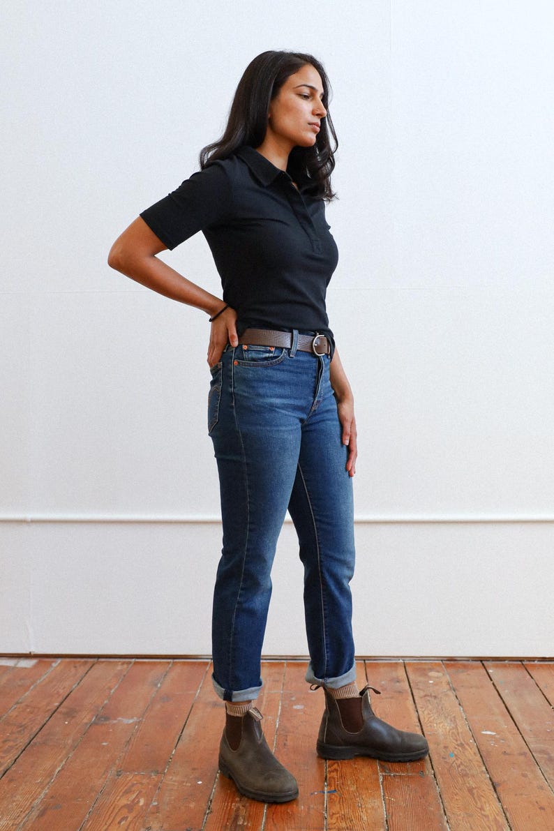 Pode incluir: Uma pessoa vestindo uma camisa preta de colarinho, jeans azul e botas de tornozelo marrons. A pessoa est&aacute; em p&eacute; em um piso de madeira.