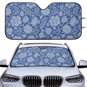 Puede incluir: Un parasol para coche azul con un patrón floral blanco. El parasol está hecho de un material ligero y está diseñado para bloquear los rayos del sol.