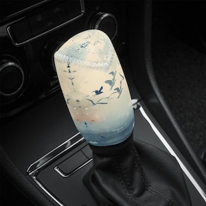 May include: A car gear stick cover featuring a scenic design. The cover is mainly white with a blue and orange gradient at the base, showing a sky and ocean scene with flying birds. The cover is cylindrical and made of a soft material.