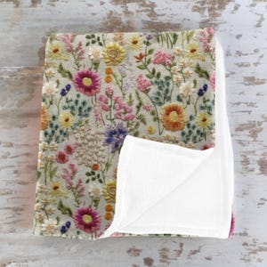 May include: A soft, plush throw blanket with a floral pattern. The front side features a white background with embroidered flowers in shades of pink, yellow, orange, and green. The back side is a solid white fleece.