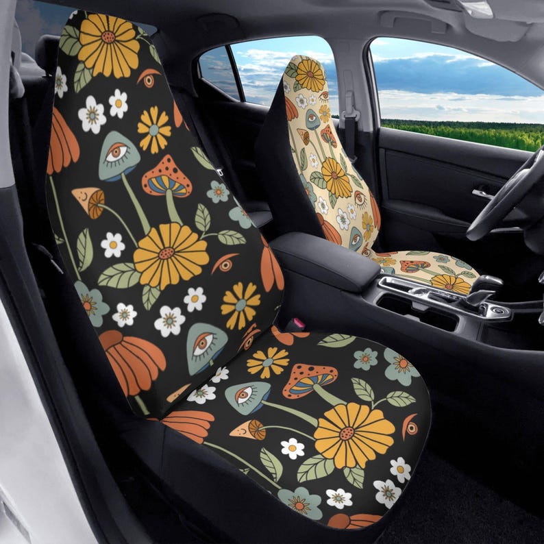 May include: A pair of car seat covers featuring a black background with a repeating pattern of yellow daisies, orange flowers, light blue mushrooms, and stylized eyes. The passenger seat cover features a cream background with the same pattern.