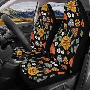 May include: A pair of car seat covers featuring a black background with a repeating pattern of yellow flowers, white blossoms, green leaves, and mushrooms with eyes. The design includes orange and blue accents throughout the fabric surface.