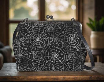 May include: Black handbag with a silver spiderweb pattern. The bag has a clasp closure and a long, adjustable strap. The bag is sitting on a wooden table. The bag is a stylish accessory.