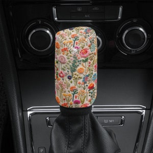 Può includere: Un copri pomello del cambio marcia bianco con un motivo di ricamo floreale colorato. Il copri pomello è in tessuto e ha un elastico nero in fondo.