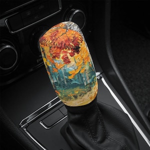 Może przedstawiać: Osłona dźwigni zmiany biegów do samochodu z motywem jesiennego krajobrazu. Osłona przedstawia żywy obraz drzew z pomarańczowymi, żółtymi i czerwonymi liśćmi. Grafika osadzona jest na tle niebieskich i zielonych odcieni, z czarnym szwem.