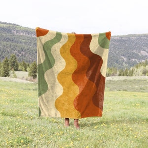 May include: A large throw blanket with a wavy pattern in shades of green, yellow, orange, and brown. The blanket is being held up by a person in a field.