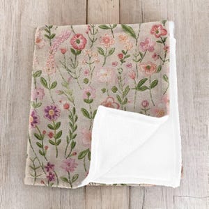 Op de afbeelding: Een zachte, pluche deken met een bloemenprint aan de ene kant en wit fleece aan de andere kant. De bloemenprint heeft roze, paarse en groene bloemen op een beige achtergrond.