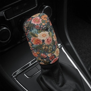 May include: A floral car gear stick cover. The cover has a dark background with a pattern of colourful flowers in shades of pink, orange, and cream. The cover is stitched with black thread.