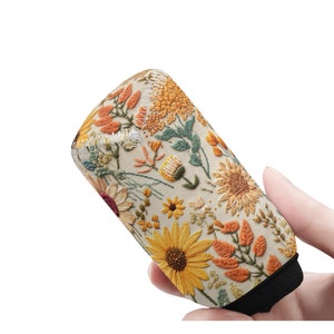 Puede incluir: Un enfriador de latas beige con un patrón floral bordado que presenta girasoles, hojas y otras flores en tonos de amarillo, naranja y verde.