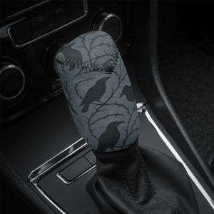May include: A grey shift knob cover with a pattern of black ravens on thorny branches. The cover is stitched at the top and fits over a car's gear shift.