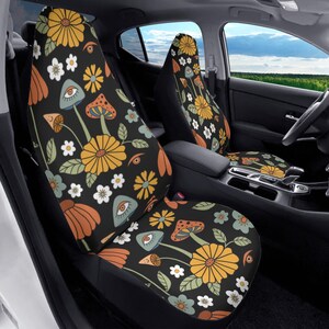 May include: A pair of car seat covers featuring a black background with a repeating pattern of yellow flowers, white daisies, orange petals, green leaves, and light blue mushrooms with human eyes integrated into the mushroom caps.