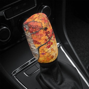 Może przedstawiać: Osłona gałki zmiany biegów z żywym wzorem liści jesiennych. Osłona ma mieszankę odcieni pomarańczowego, żółtego i czerwonego, z czarnym szwem. Projekt przypomina malowanie jesiennych liści.