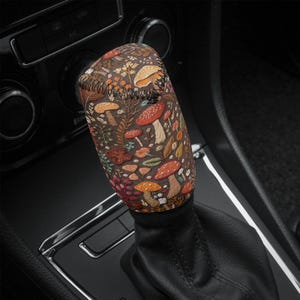 May include: A shift knob cover with a vibrant mushroom and foliage design on a dark brown backdrop. The fabric cover fits over a car's gear shift lever. The pattern includes different mushrooms and leaves, adding a touch of nature to the car's interior.