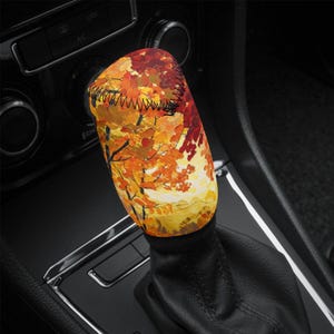 Puede incluir: Una colorida funda para el pomo del cambio con un diseño de paisaje otoñal. La funda muestra una pintura vibrante de follaje otoñal en tonos rojos, naranjas y amarillos, con costuras negras en la parte superior. La funda está en una palanca de cambios negra.