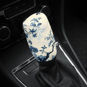 May include: A cream-colored shift knob cover with a blue floral pattern. The cover is cylindrical and fits over the gear shift of a car. The design features stylized flowers and branches.