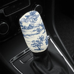May include: A car gear stick knob with a blue and white toile design. The pattern depicts a rural scene with trees, a house, and a fence. The knob is cylindrical and sits on a black leather gear stick gaiter.