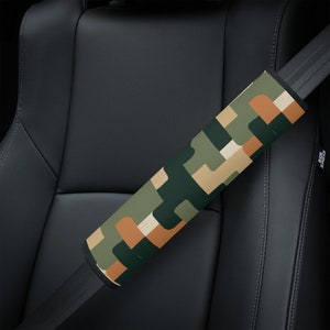 May include: A patterned seat belt cover featuring a camouflage design in green, tan, and brown hues. The cover is cylindrical and designed to wrap around a seat belt. The background shows a black car seat.
